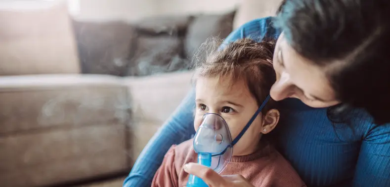 A pediatric specialty pharmacy patient wears an oxygen mask and sits in her mother's lap