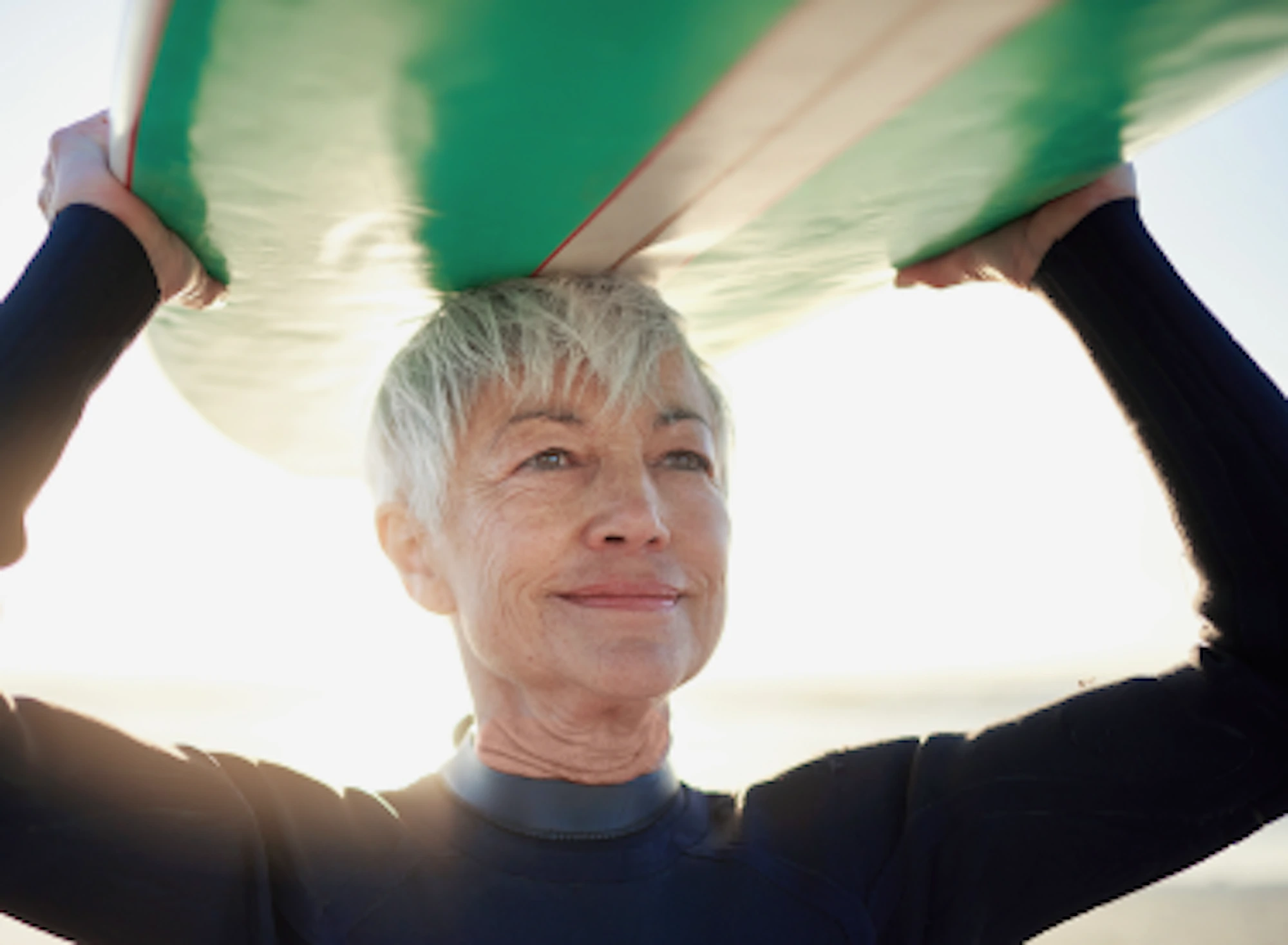 A woman with silver hair carrying her surfboard over her head while standing on a beach