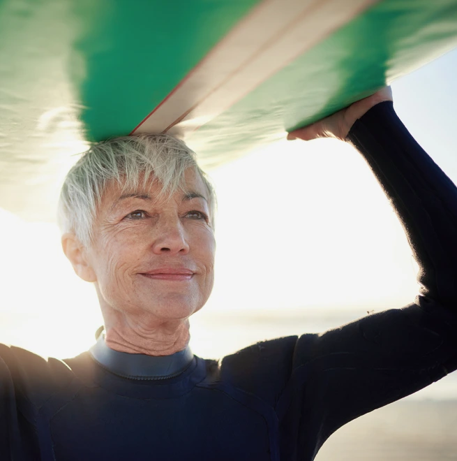 A woman with silver hair carrying her surfboard over her head while standing on a beach